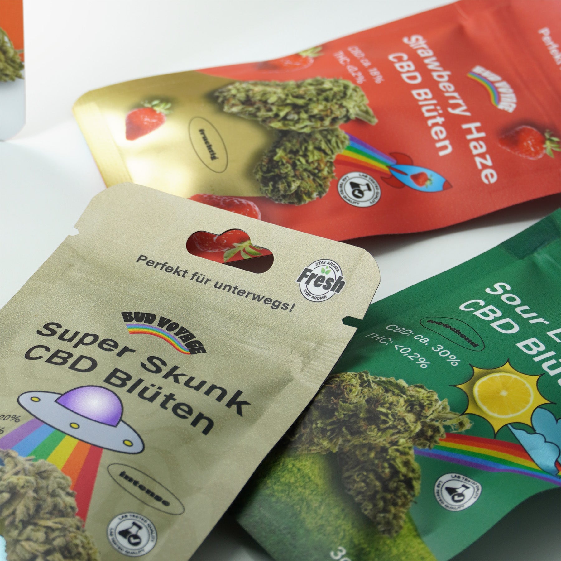 Produktfotografie:  Drei  CBD Blüten-Verpackungen zeigen Super Skunk mit UFO und Regenbogen, Strawberry Haze mit Erdbeeren und  Sour Lemon mit Zitrone, alle mit  Labortestsiegeln und  bunt
